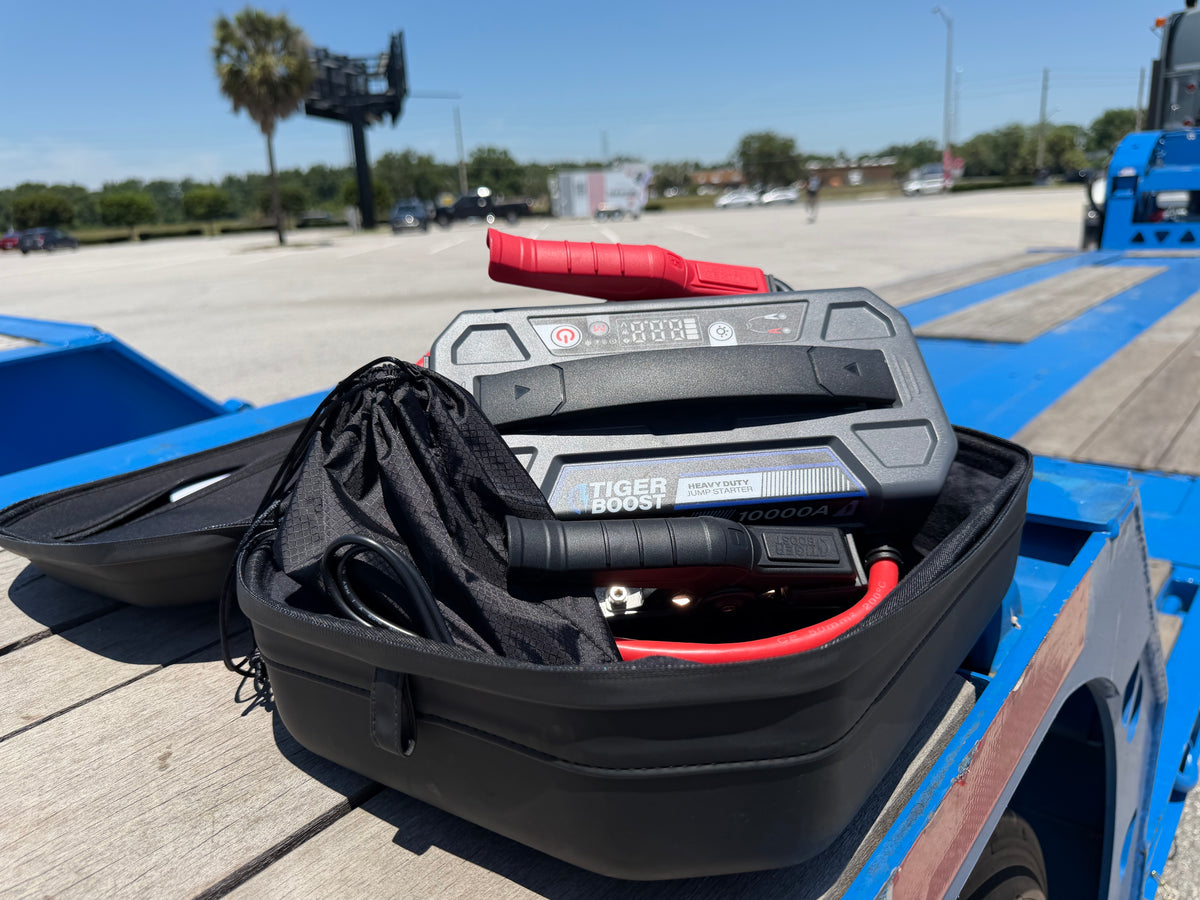 A TigerBoost portable jump starter in an open black hard case, placed on the wooden deck of a blue flatbed trailer.